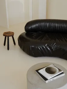 A minimalist black leather sofa in a living room setting, next to a vintage wooden stool and a concrete table with books.