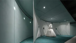 A wedding dress displayed on a mannequin within a cluttered space with white fabric curtains and green carpeting.