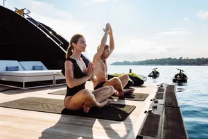 A couple practicing minimalist yoga on a boat deck near the water with a luxury yacht in the background.