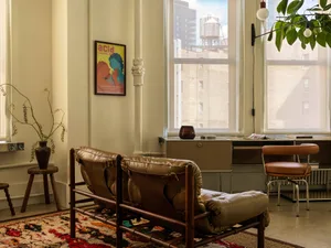 A modern living room with a leather and wood couch, a wooden stool with a vase, a wooden floor with an orange and white rug,