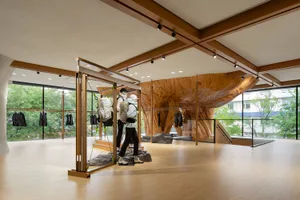 A modern retail storefront display featuring a large wooden tree trunk and mannequins with backpacks, set against a glass wal