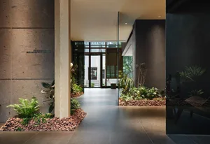 A modern lobby entrance featuring concrete walls, glass partitions, wooden columns, and lush greenery.