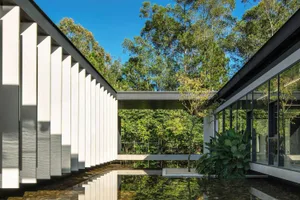 Modern house with white walls and glass windows surrounded by green trees. Concrete pillars and a small pond in the foregroun