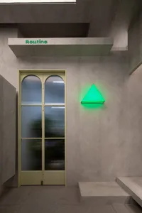 A minimalist retail interior with concrete walls and a green neon triangle light on a textured concrete wall next to a glass 