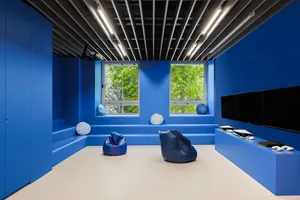 A modern industrial game room with blue seating and bean bags, featuring a television and greenery outside the windows.