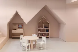 A minimalist children's playroom with wooden furniture, including a small table, chairs, a kitchenette, and a bookshelf.