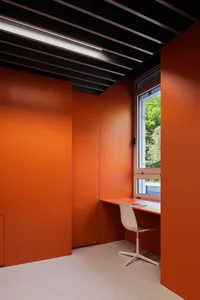 A modern home office featuring orange walls and a white chair in front of a window with greenery outside.
