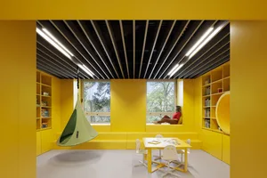 A modern playroom with yellow walls and furniture, featuring a green hanging chair, a white table with chairs, and a window o