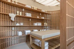 A serene minimalist spa room with wooden shelves and plush seating, featuring neatly folded white towels and candles.