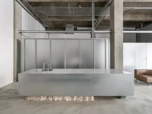 A modern kitchen with a large grey counter and industrial-style office space in the background, featuring exposed concrete wa
