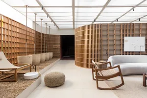 A modern living room with wooden walls and furniture, featuring a white lounge chair, a round pouf, and a curved wooden sofa.
