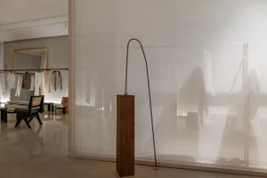 Modern minimalist room divided by sheer white curtains, featuring a sleek black sculpture on a wooden pedestal and minimalist