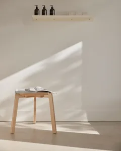 A minimalist white room featuring a wooden stool with a book on top, casting shadows on the wall.