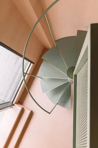 A modern metal staircase with pastel-colored steps and railings inside a room with peach-colored walls and a window with whit