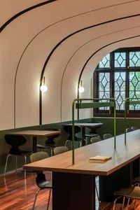 A minimalist cafe interior featuring a long wooden table with green dividers and a window with stained glass.
