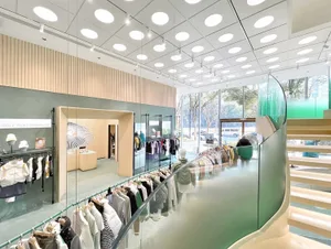 現代零售店室內，展示服飾陳列於衣架與層架上，搭配玻璃樓梯與大型窗戶。