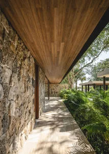 A modern walkway with stone walls and a wooden ceiling and floor, surrounded by lush greenery.