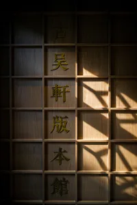 A wooden bookshelf with Japanese characters in gold standing out against a dark background with sunlight filtering through.