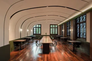 A modern cafeteria with wooden tables and chairs, featuring large windows and a curved ceiling with recessed lighting.