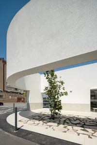 A minimalist white concrete building with a curved facade and a tree planted in a stone garden on a sunny day.