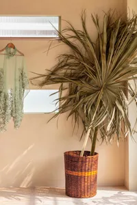 A woven basket with dried palm fronds on a beige wall next to a hanging piece of embroidered fabric.