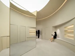 A minimalist wardrobe room featuring concrete walls and flooring, with a person sitting and reading on the curved bench.