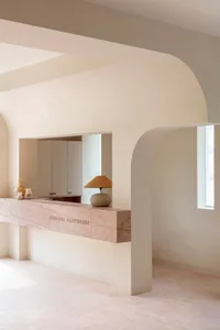 A minimalist interior space with neutral tones, featuring a wooden counter with a lamp and decorative items, and an arched do