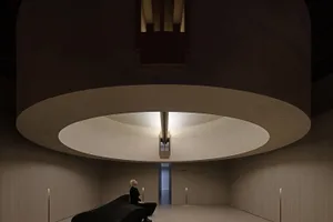 A minimalist interior with a white concrete floor and ceiling, featuring a circular light fixture and a woman sitting on a bl