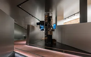 A modern installation featuring a vertical stack of wooden boxes illuminated by blue light in a gallery setting.