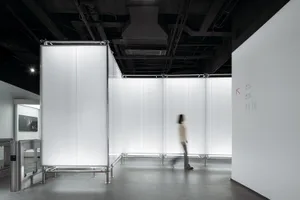 A minimalist exhibition space featuring glass walls and a single blurred person walking through the space.