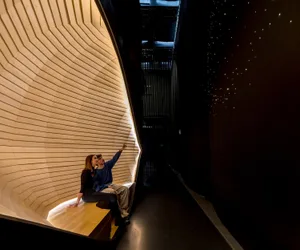 Two people sitting in an indoor space with a wooden, angular design, illuminated by warm lighting.