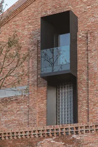 A modern building with a facade made of red bricks. A glass window with black frame and a glass balcony are visible.
