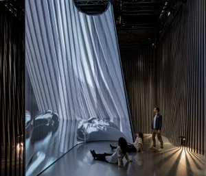 People observing a modern art installation featuring a light-structure on a mirrored floor surrounded by vertical lines.