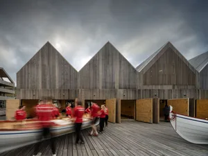 People in red uniforms moving a boat on a wooden deck with a wooden building in the background.