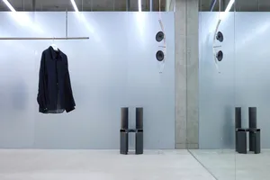 A minimalist furniture piece made of black metal against a white background, with a shirt hanging in the background.