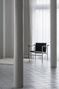 Minimalist design featuring a black metal and leather armchair in a bright, modern room with white flooring and curtains.