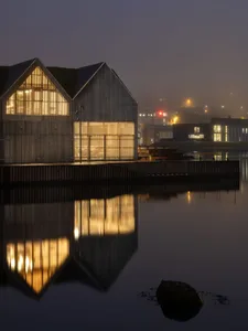 Modern wooden building with illuminated windows reflected in calm water at night.