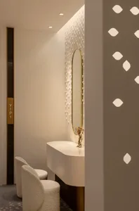 A modern hotel suite with white furniture, a vanity area with an oval mirror and decorative lighting, and a textured carpet.