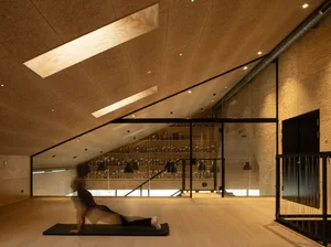 A person in a minimalist fitness studio practicing yoga on a wooden floor with a glass wall showcasing trophies and awards.