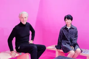 Two people sitting on benches with a pink background, dressed in black clothing.