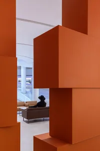 A modern lobby with large, orange, geometric structures and a woman wearing a black hat sitting on a brown sofa.
