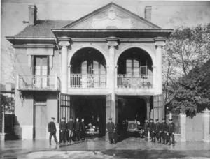 Black and white photograph of a historical fire station exterior with multiple firefighters in uniform standing in front of t