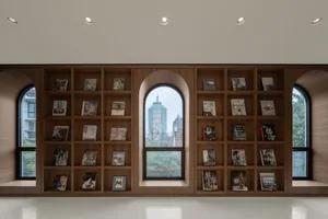 A library bookstore with wooden shelves filled with books and framed pictures, featuring large windows with a view of urban b