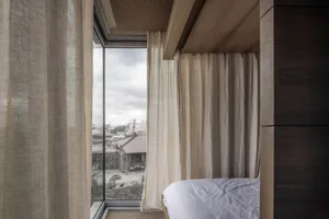 A minimalist bedroom with floor-to-ceiling curtains and a view of the surrounding rooftops and clouds.