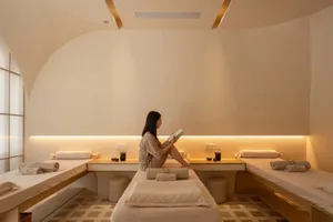 A woman wearing a beige robe sits on a spa bed in a minimalist spa room with beige fabric and warm lighting.