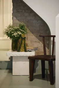 A rustic wooden chair sits beside a white marble side table adorned with flowers and books, against a backdrop of a stone sta