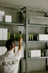A woman in an 'Ikkari' branded jacket arranging green racks in a store setting.