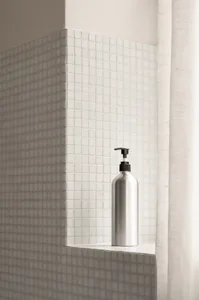 A metallic bottle with a black dispenser cap, placed on a shelf against a backdrop of white square tiles.