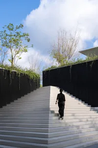 A person walking up a concrete staircase bordered by a black wooden fence adorned with greenery against a clear blue sky, emp