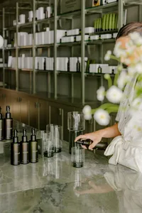 Person pouring a container of liquid into a clear glass in a spa setting with glass shelves and flower arrangements.